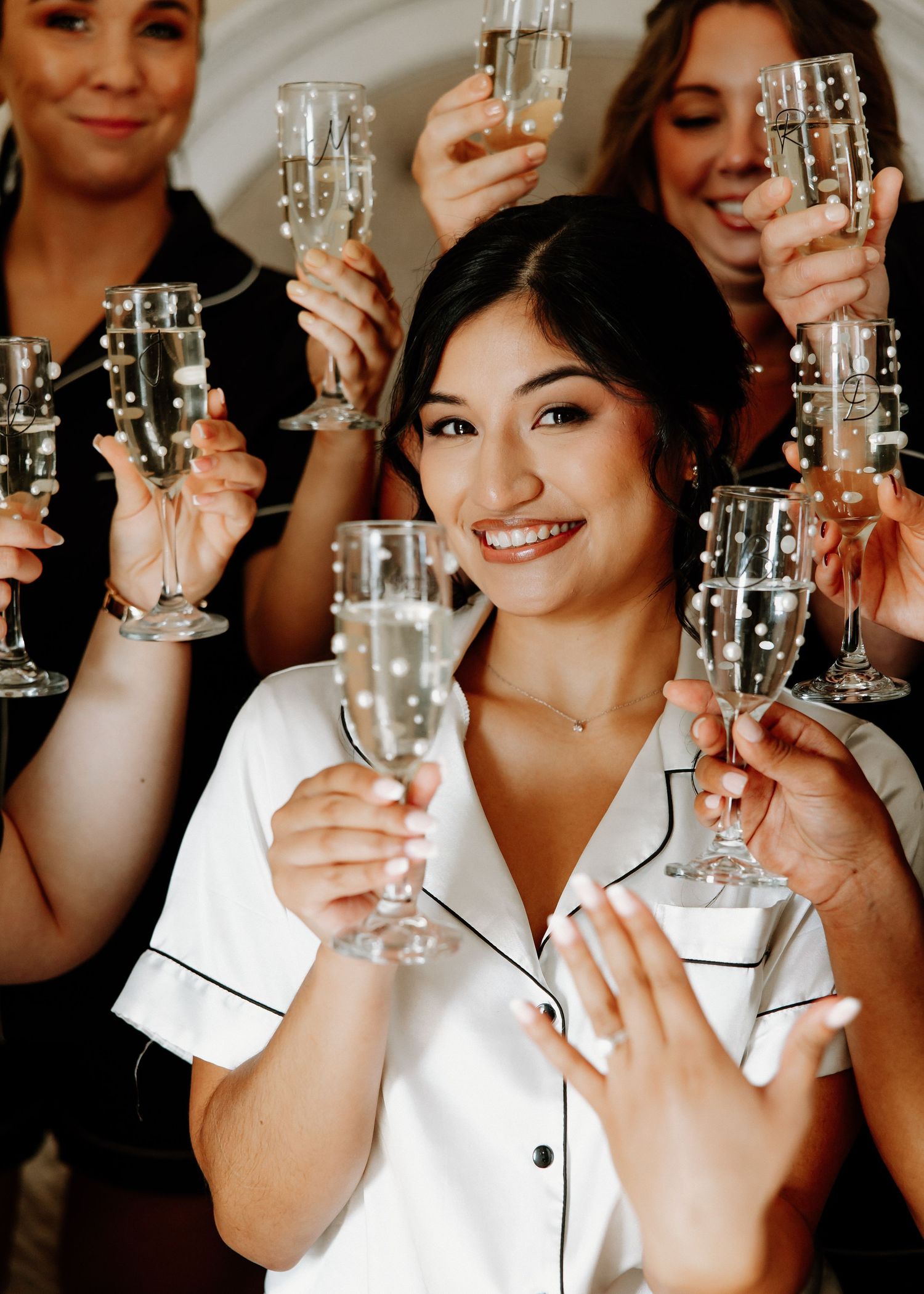 Smiling bride in white satin pajamas toasting champagne with bridesmaids during wedding morning prep.