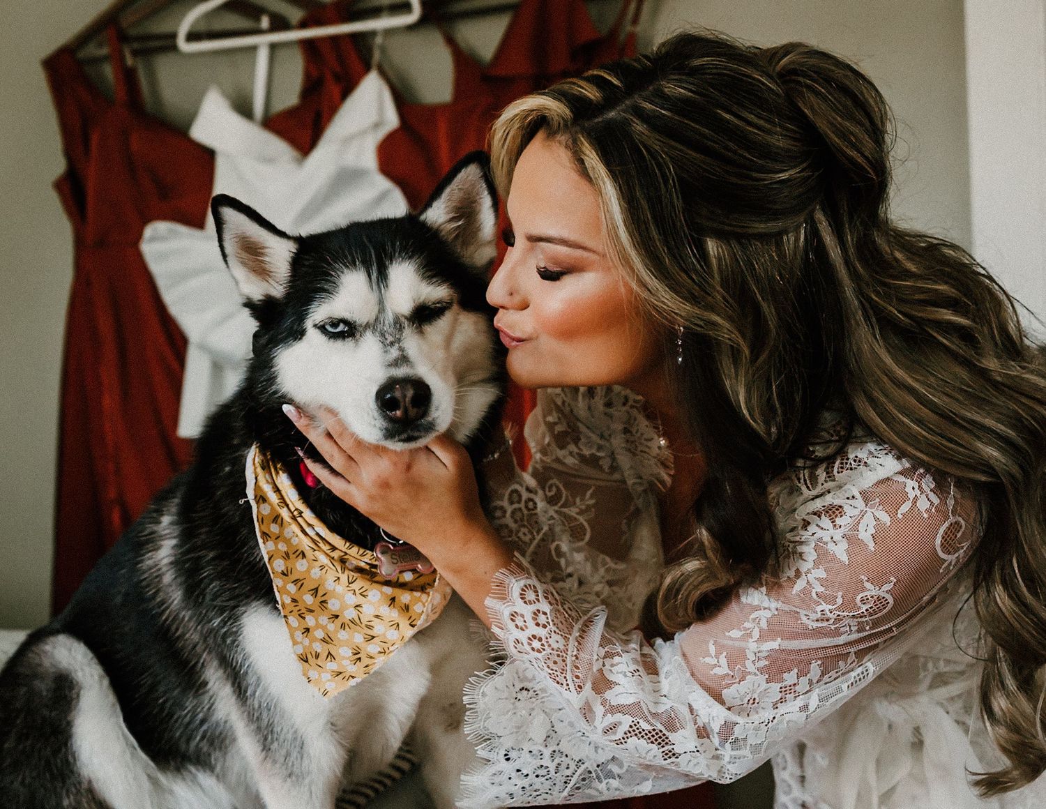 Bride in lace robe kissing her Siberian Husky dog wearing a bandana before wedding.