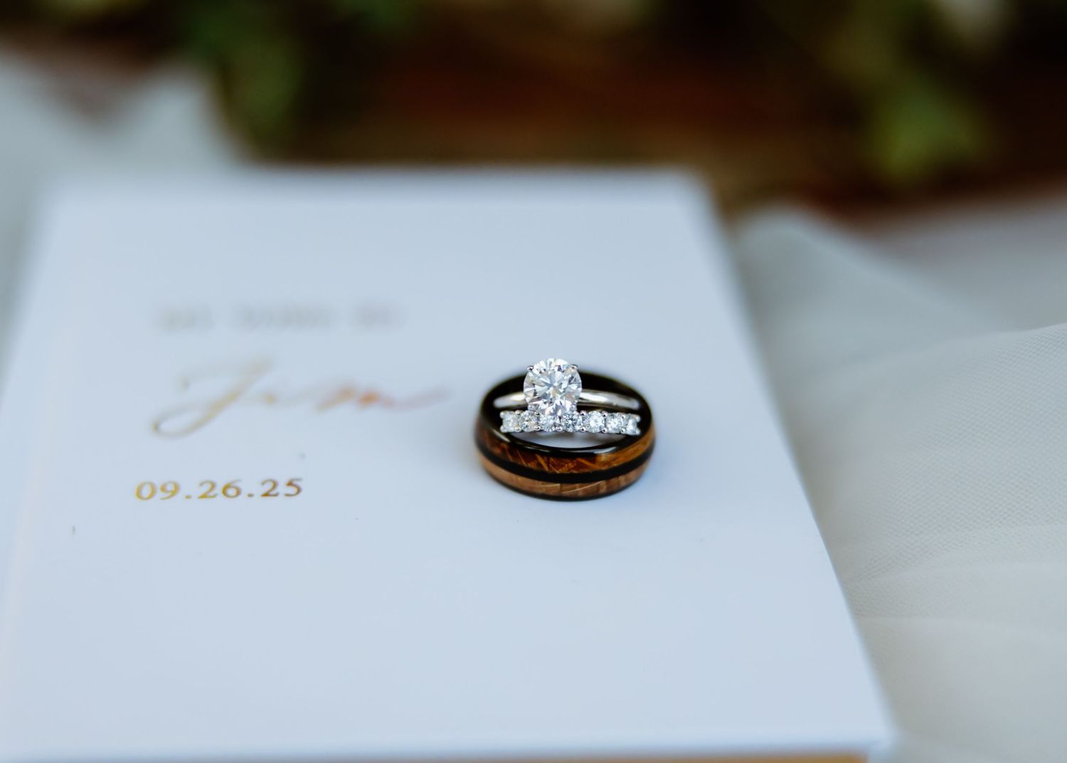 Diamond engagement ring and wooden wedding band resting on elegant white wedding vow booklet.
