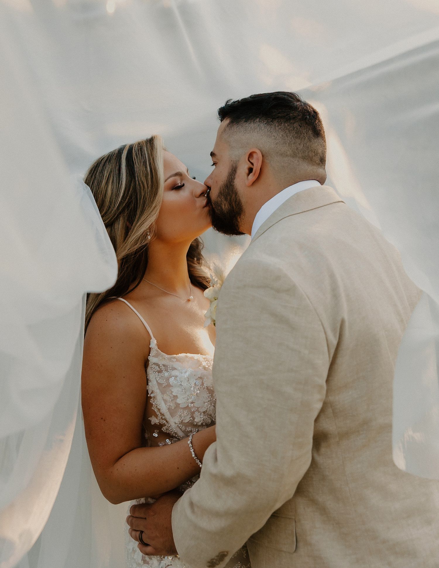 Bride and groom sharing romantic kiss under flowing white veil in warm golden light.