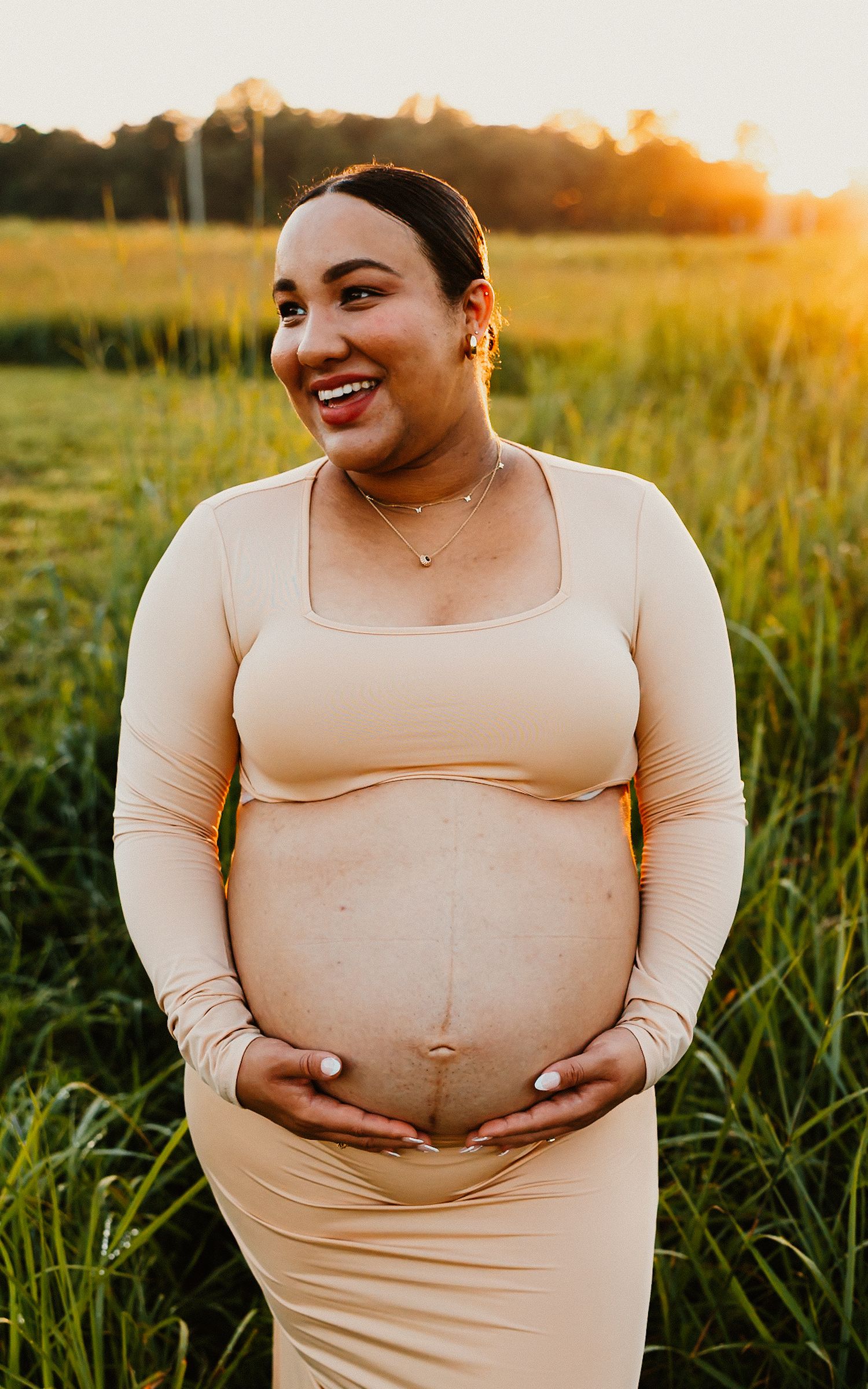 Smiling pregnant woman in beige dress cradling baby bump in sunlit green field at sunset.