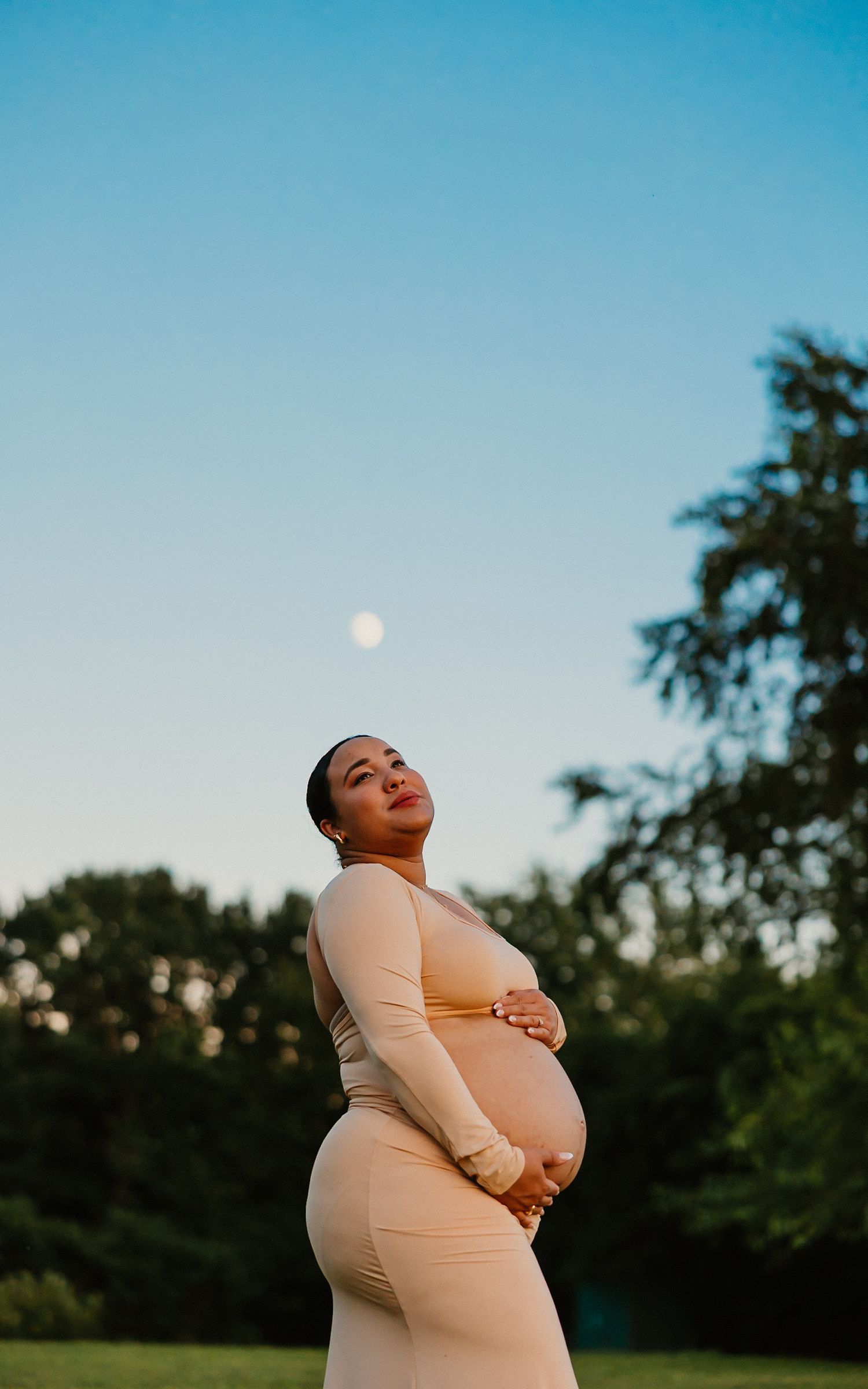 Pregnant woman in beige dress gazing upward at full moon in clear blue evening sky.