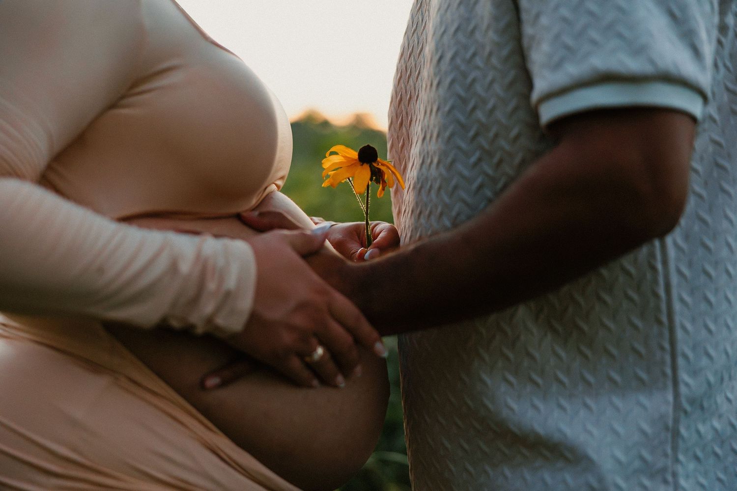 Pregnant woman in beige dress holds baby bump while partner offers a yellow flower outdoors.