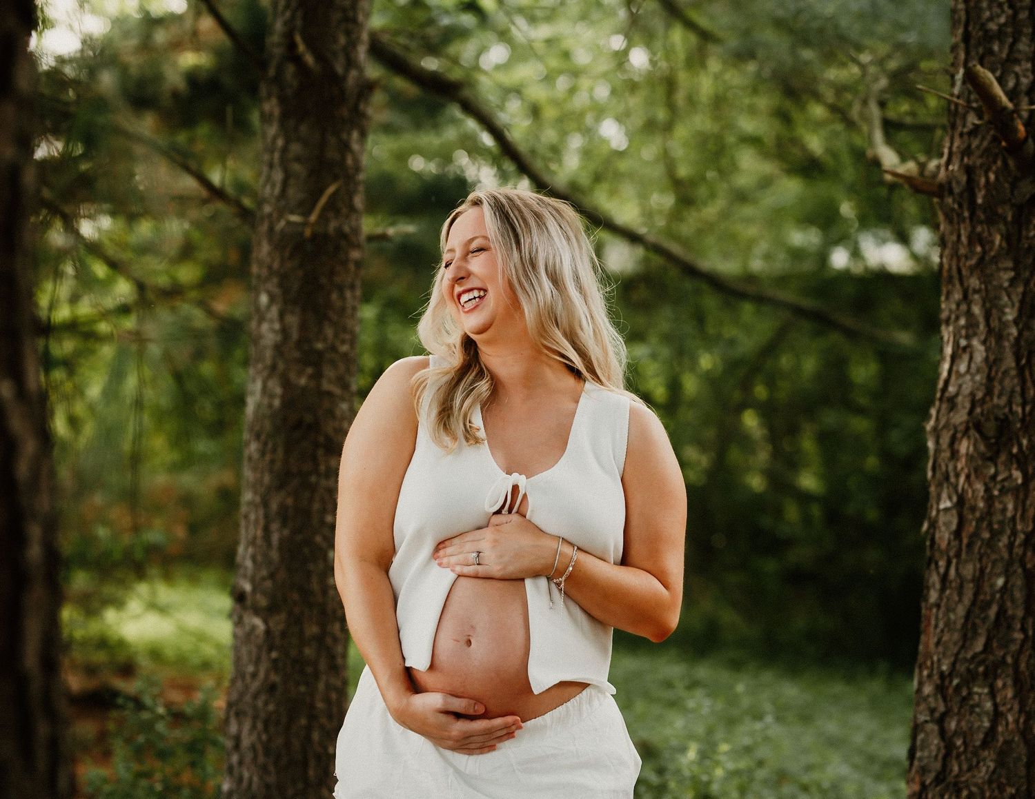 Happy pregnant woman in white outfit cradles baby bump while standing among trees outdoors.