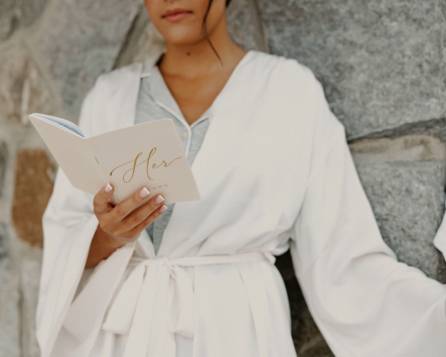 Bride in white robe reads vows from a book labeled Her before wedding ceremony.
