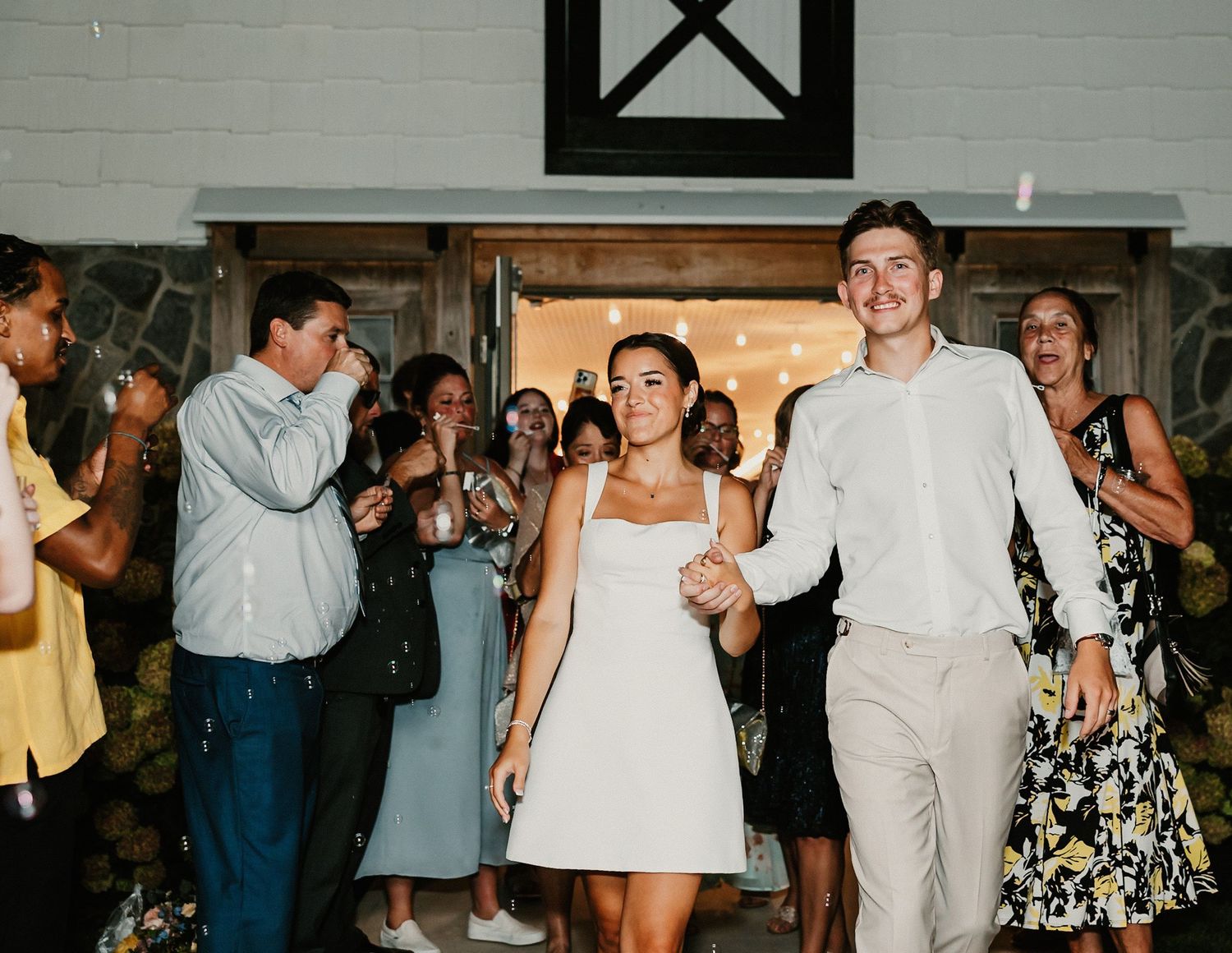 Smiling newlywed couple exits barn wedding venue as guests celebrate around them.