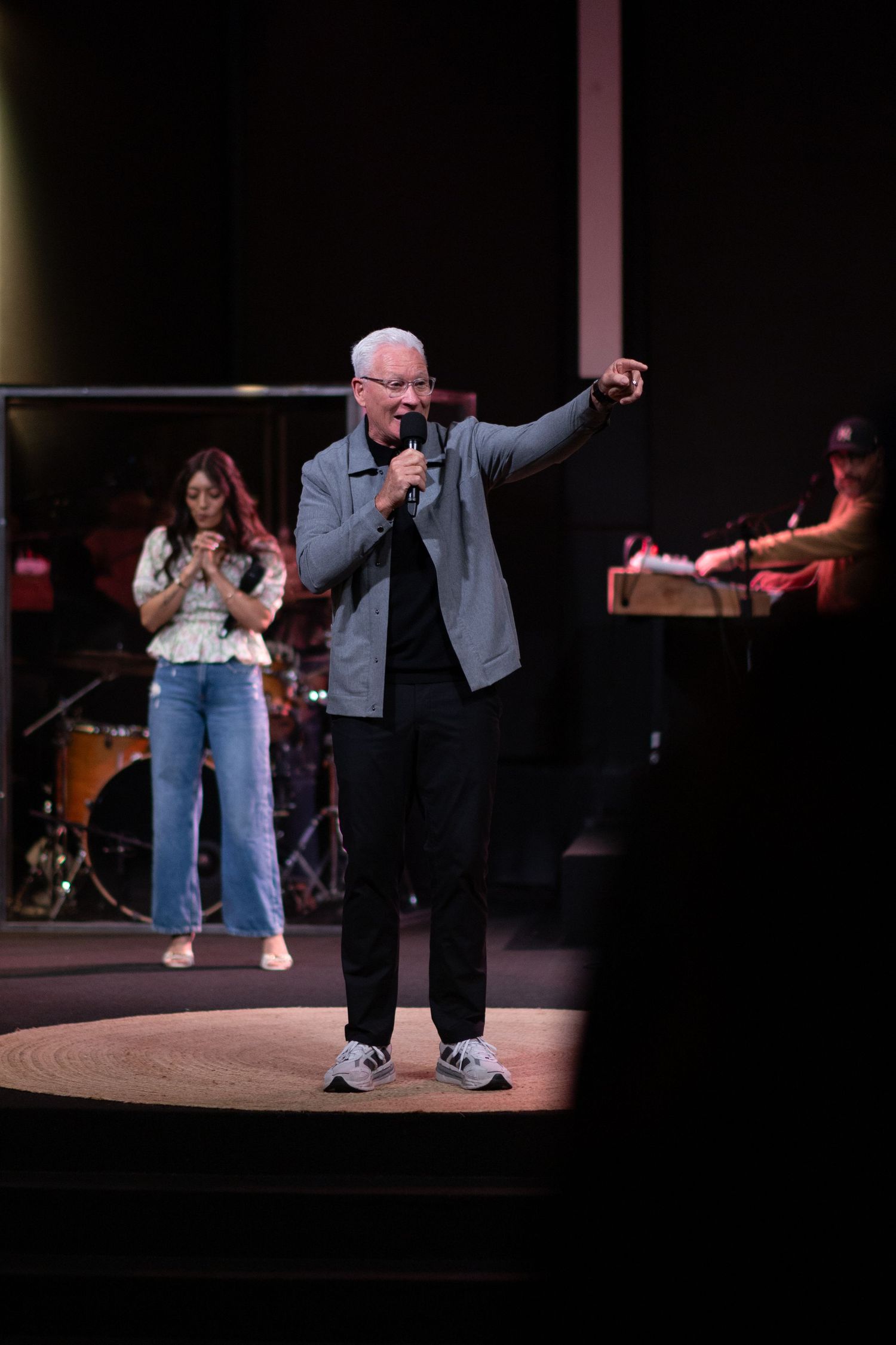 Older man with white hair speaks into microphone on stage, pointing outward, with band and singer visible behind him.