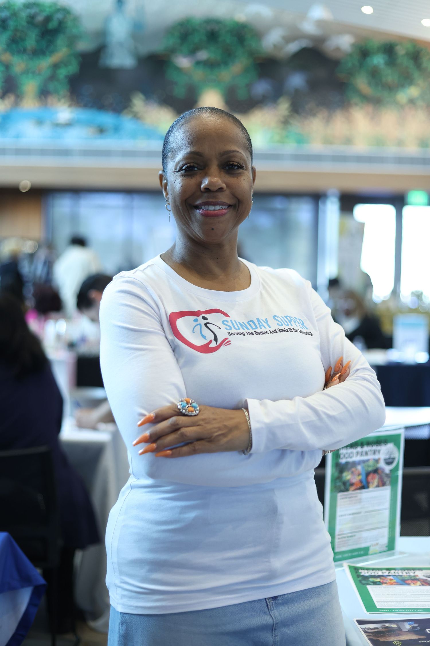 Woman in white long-sleeve shirt with logo stands smiling with arms crossed at an indoor event.