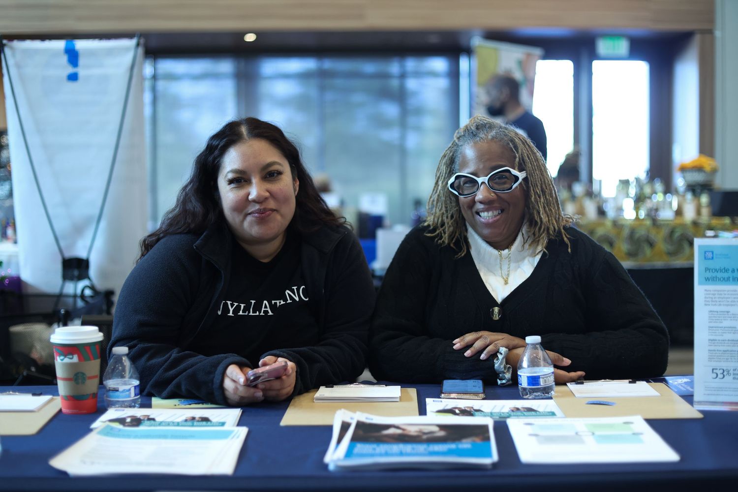 Two women smile at an informational table covered with brochures and water bottles at an indoor community event.