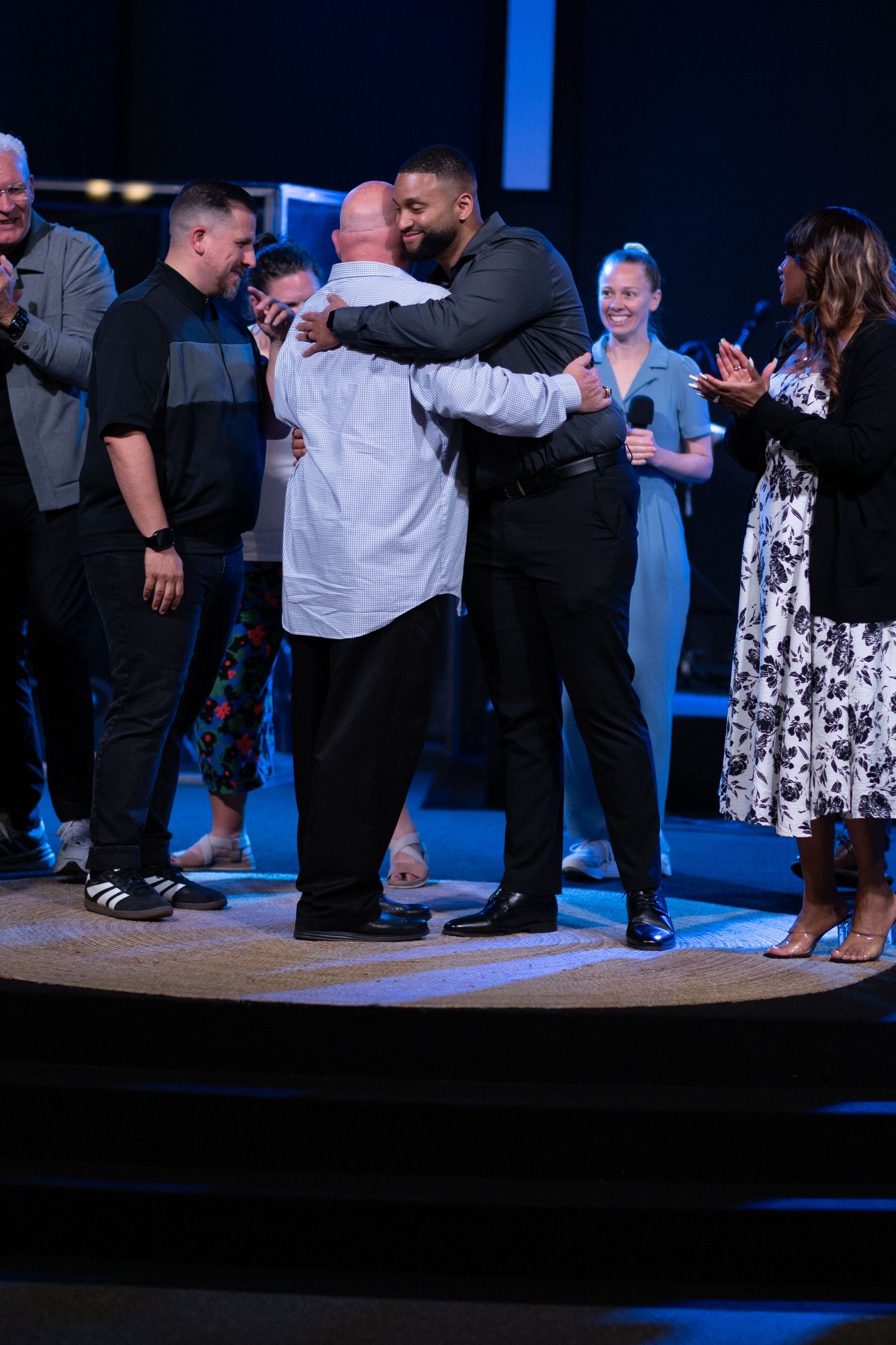 Two men sharing an emotional hug on stage while surrounded by applauding audience members.