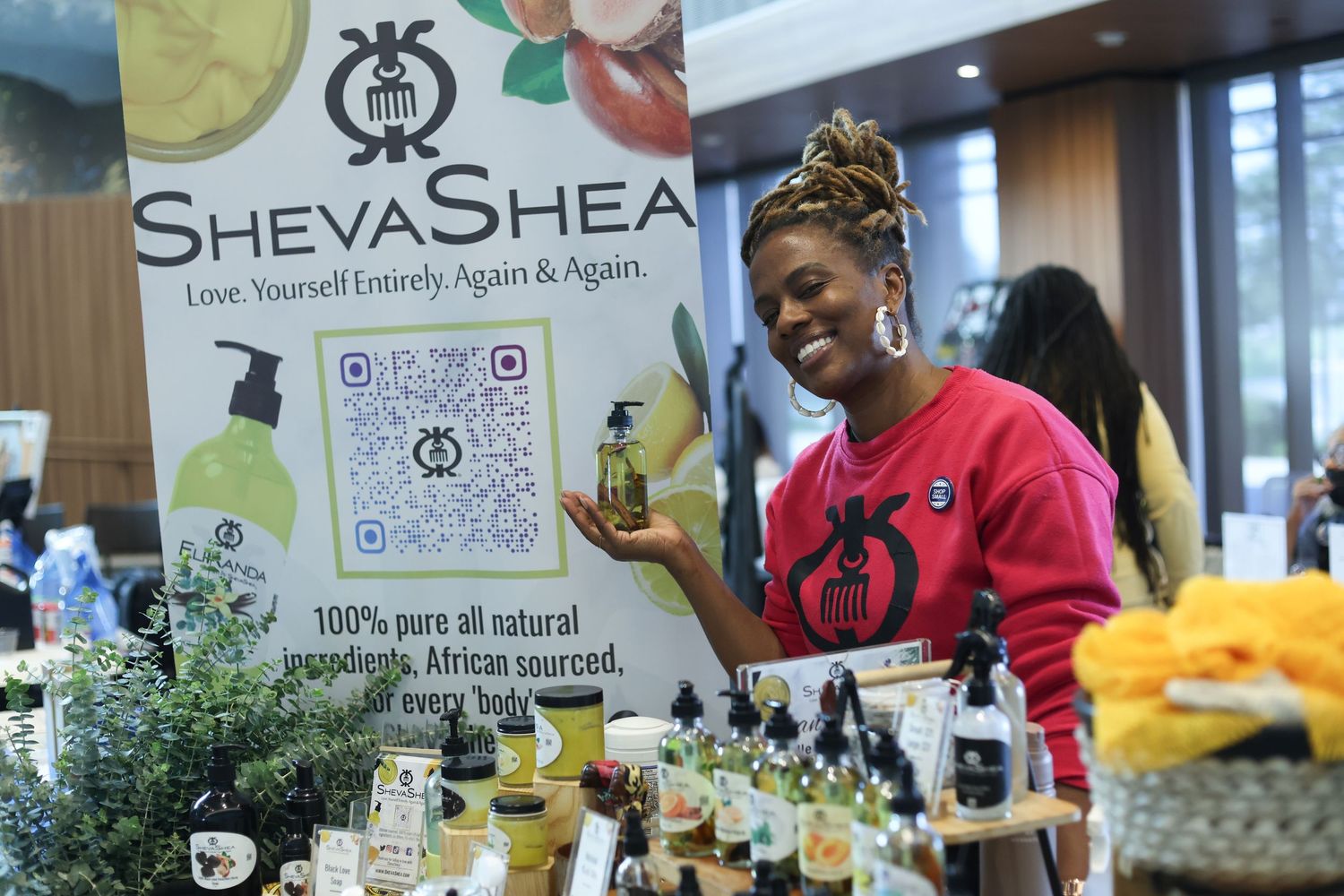 Woman smiling at a ShevaShea natural African-sourced beauty products booth displaying lotions and oils.