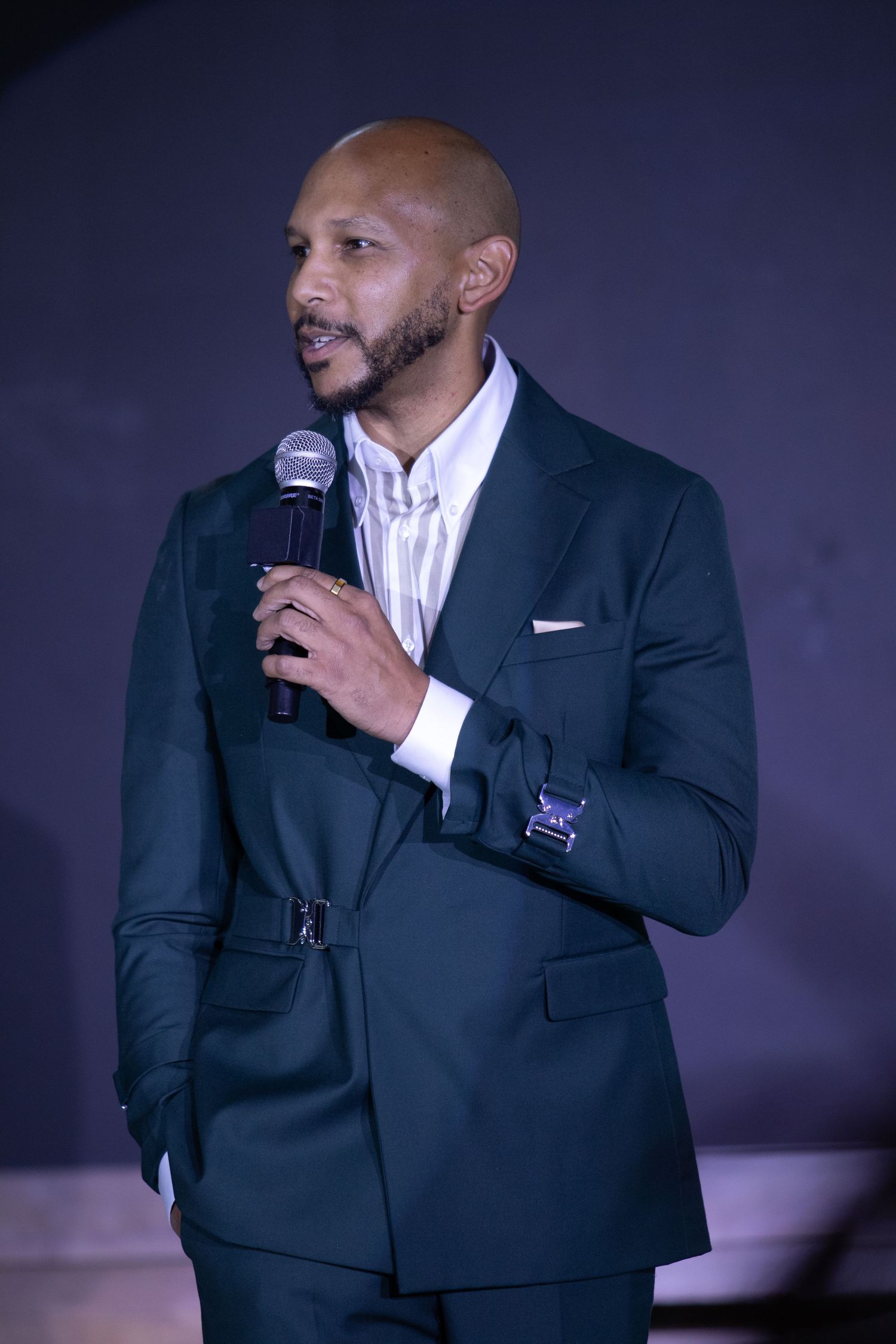Bald Black man in dark green suit speaking into microphone at formal event.