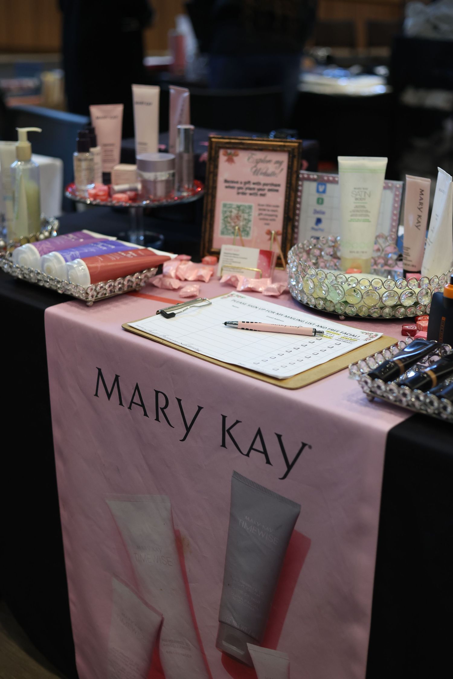 Mary Kay cosmetics display table with pink runner, beauty products, clipboard, and decorative trays at a promotional event.
