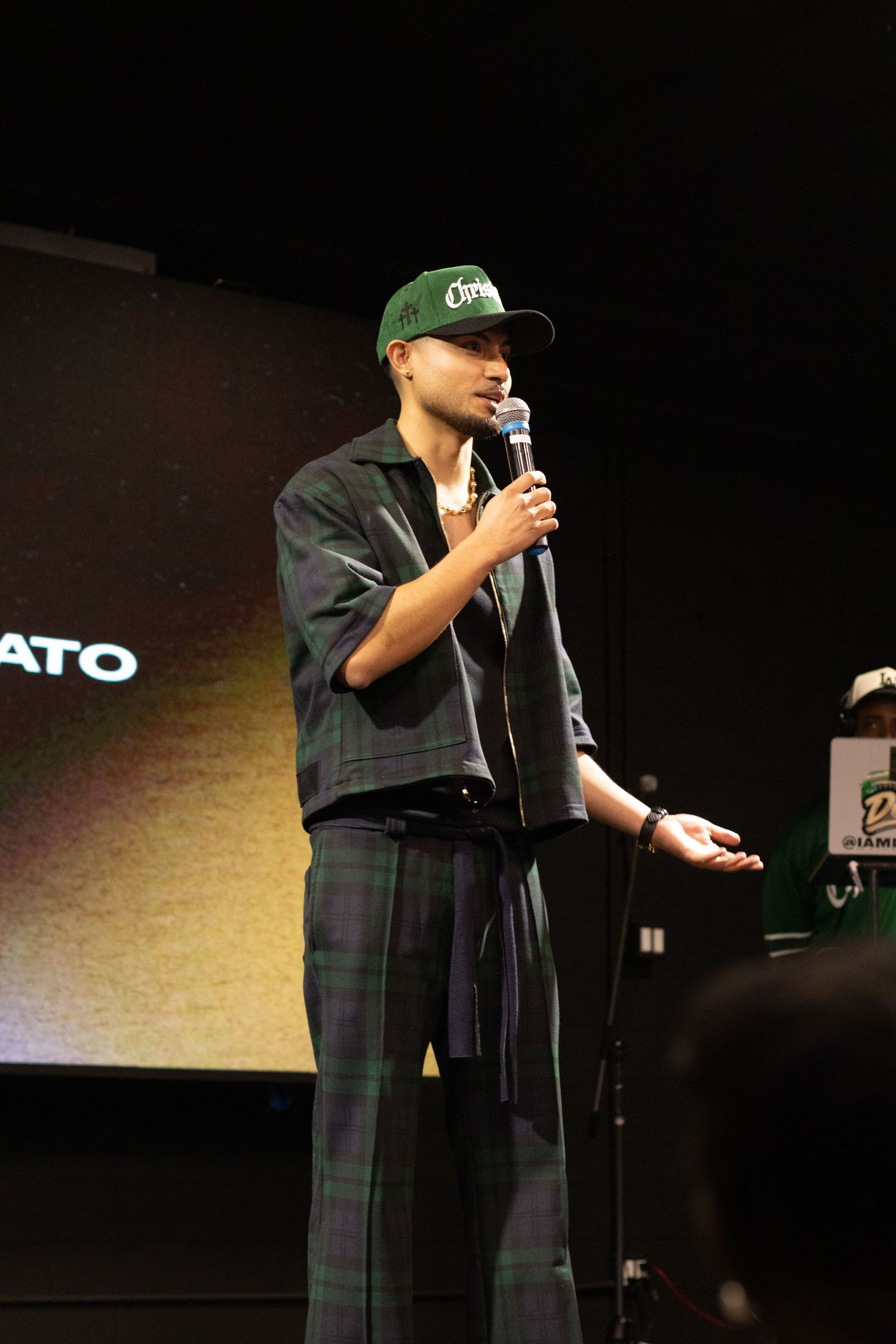Young man in green cap and plaid pants speaking at microphone on dimly lit stage.