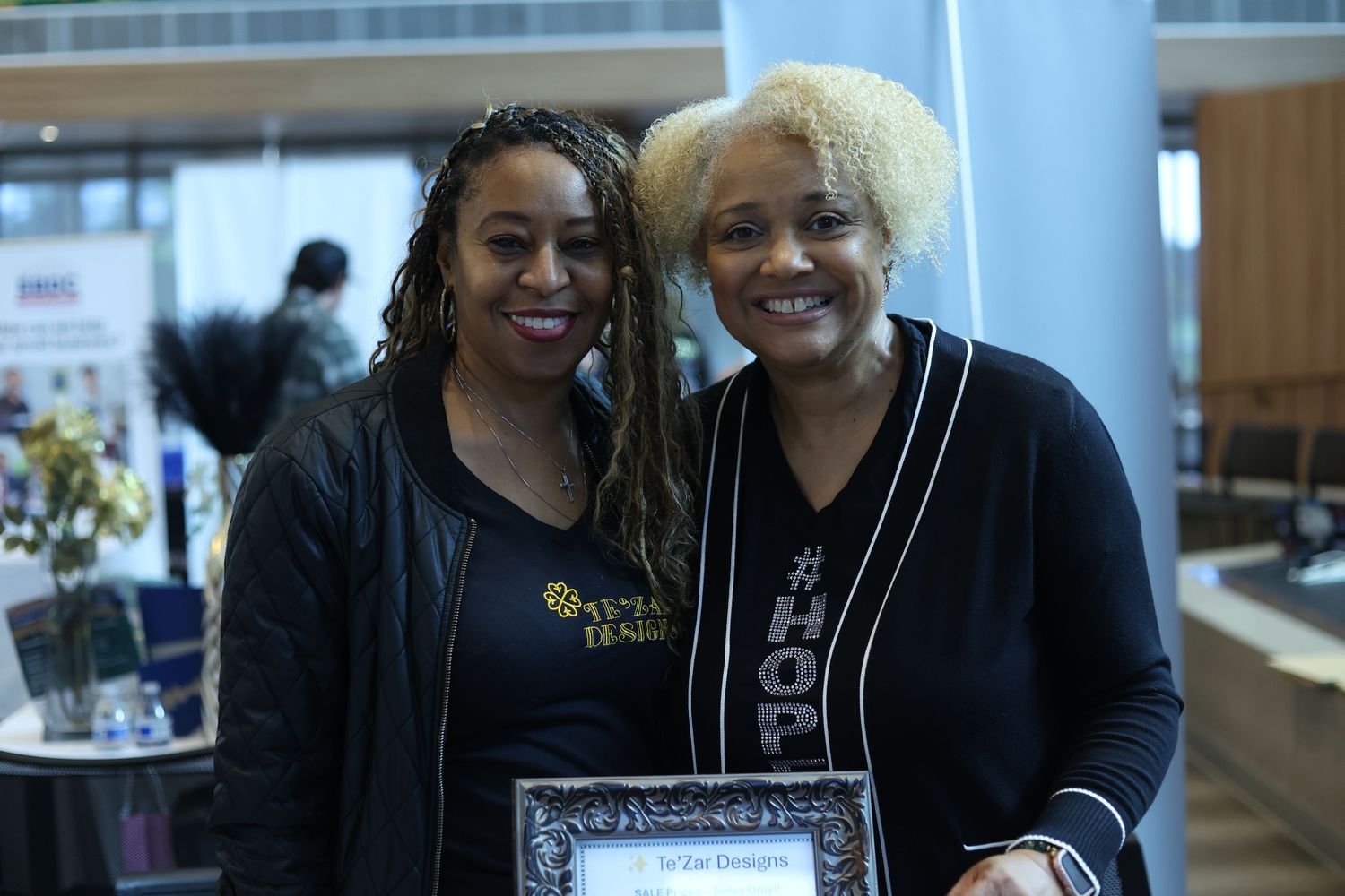 Two women smiling together at an indoor event, one holding a framed award plaque.