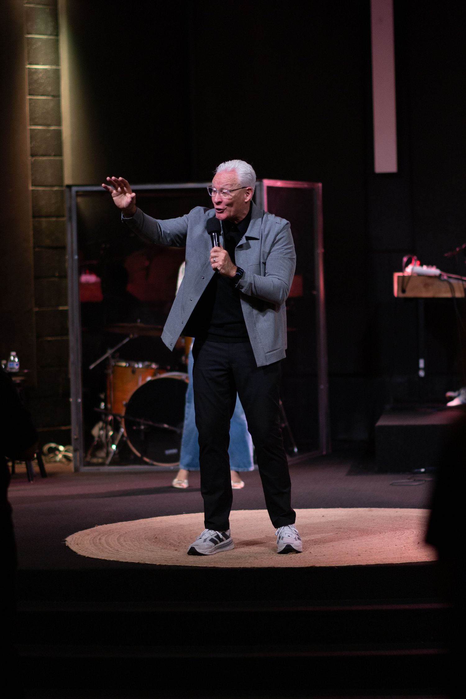 Elderly man speaking on stage with microphone, wearing gray blazer and black pants, with drum kit visible in background.