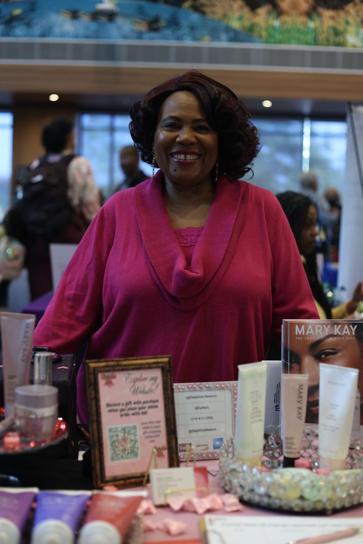 Smiling woman in pink top stands at a Mary Kay cosmetics display table at an indoor vendor event.