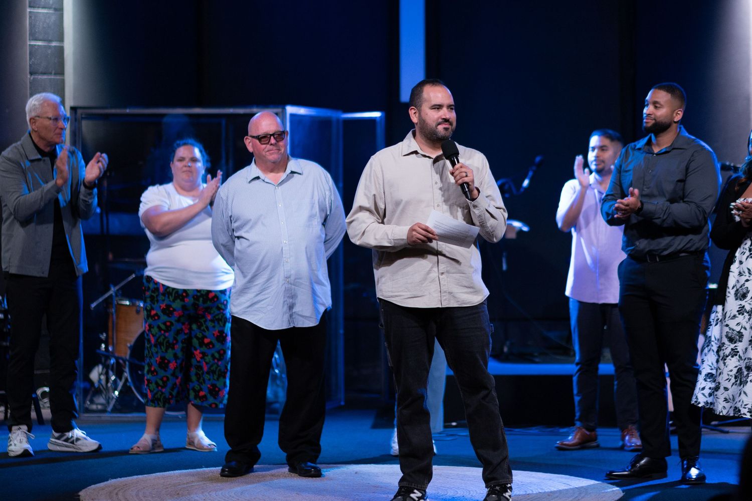 Man speaks into microphone on stage with group of people standing behind him in a modern church setting.