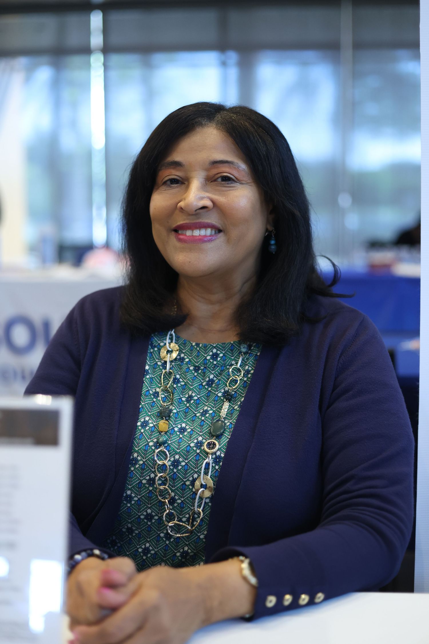 Smiling older woman with dark hair wearing a blue cardigan and patterned blouse with a long necklace, seated at a table.
