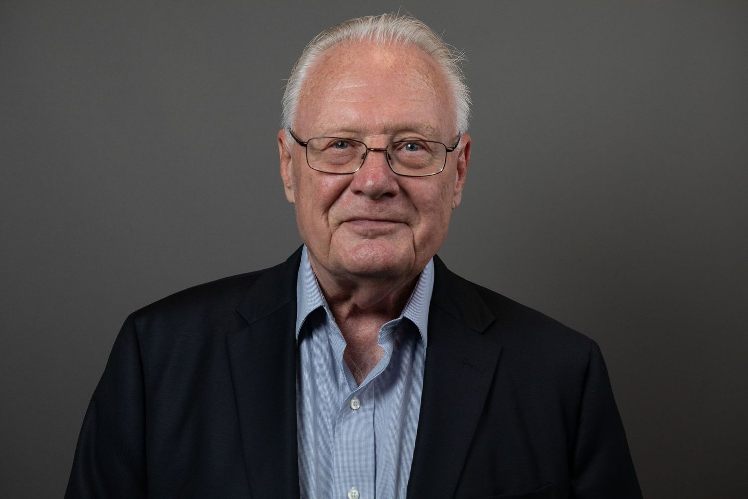 Elderly man with white hair and glasses wearing a dark blazer and light blue shirt poses for a professional portrait.