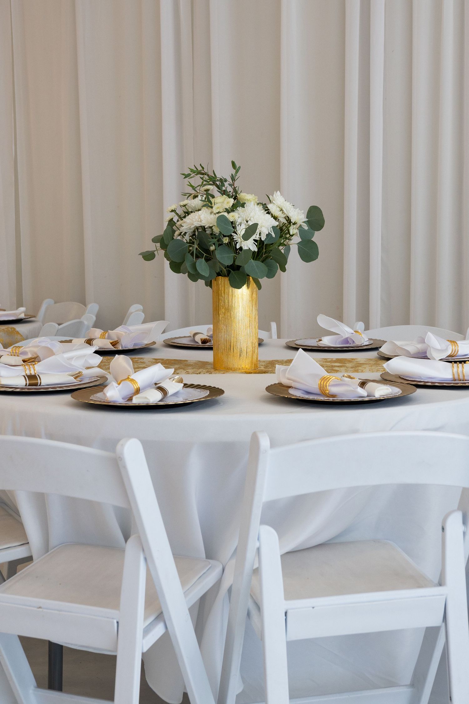 Elegant wedding reception table with white linens, gold chargers, folded napkins, and a gold vase centerpiece with white flowers.
