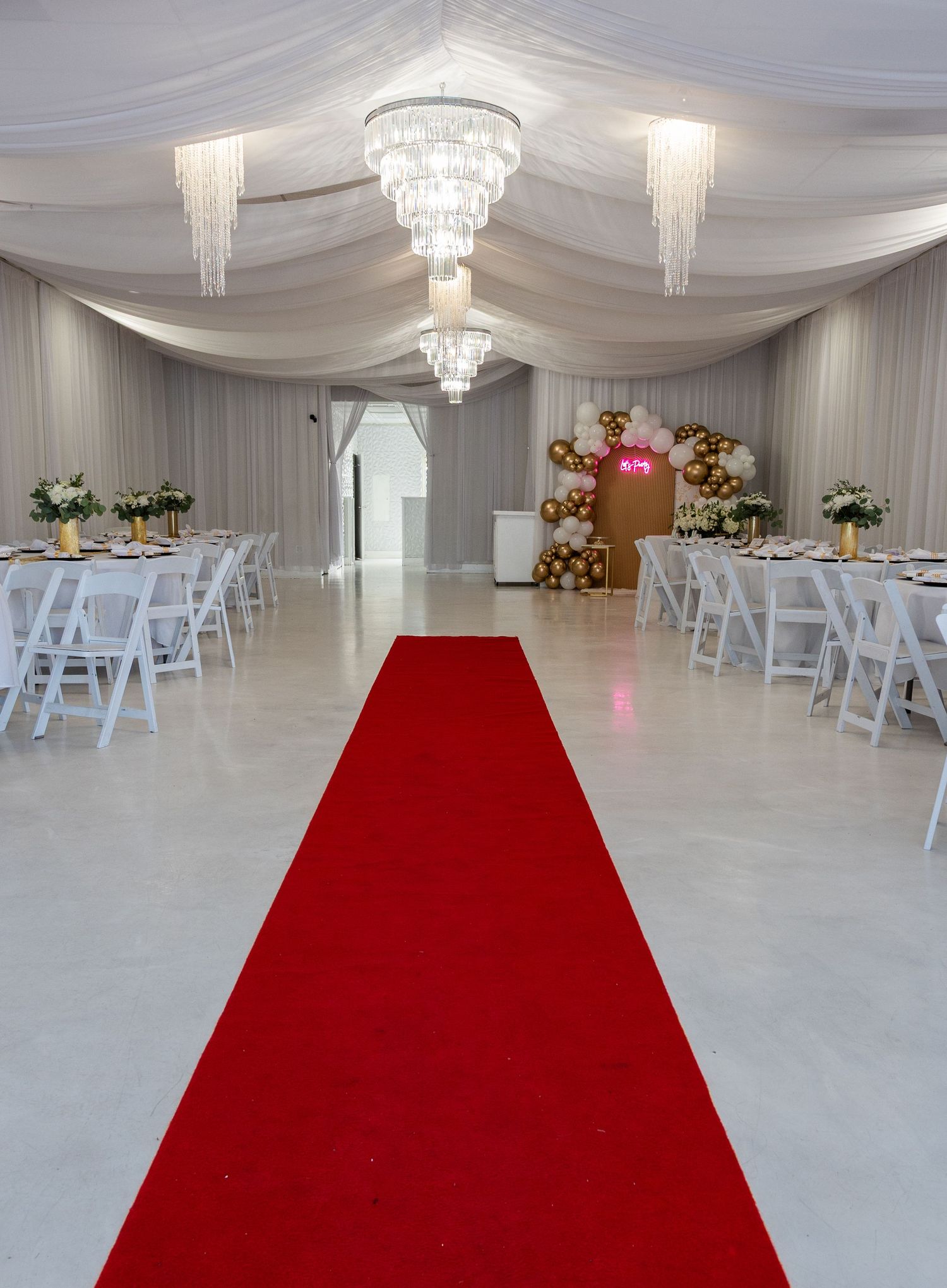 Elegant event hall with red carpet aisle, crystal chandeliers, white draping, and decorated tables for a formal celebration.