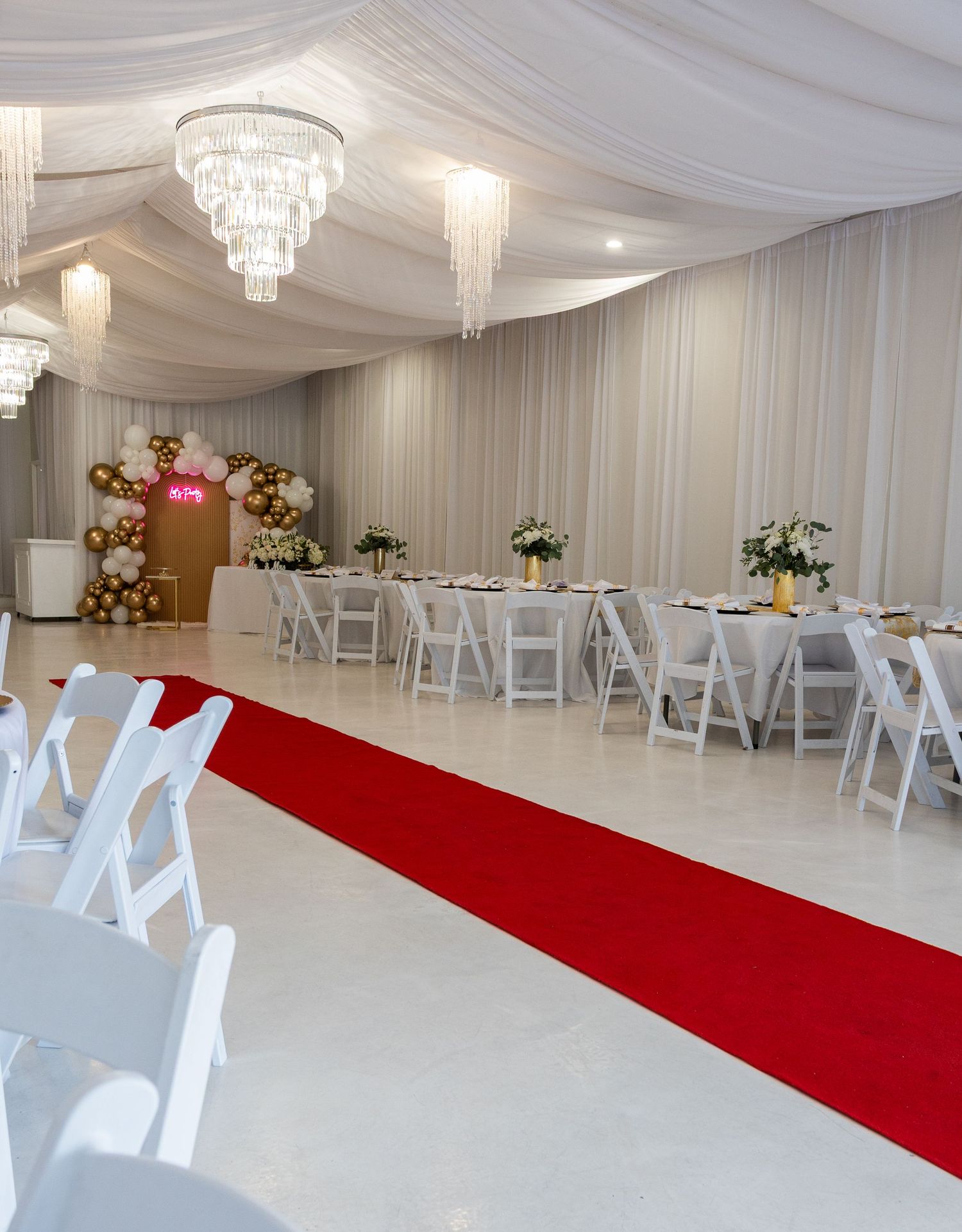 Elegant event venue with red carpet aisle, white folding chairs, draped ceilings, crystal chandeliers, and balloon arch decor.