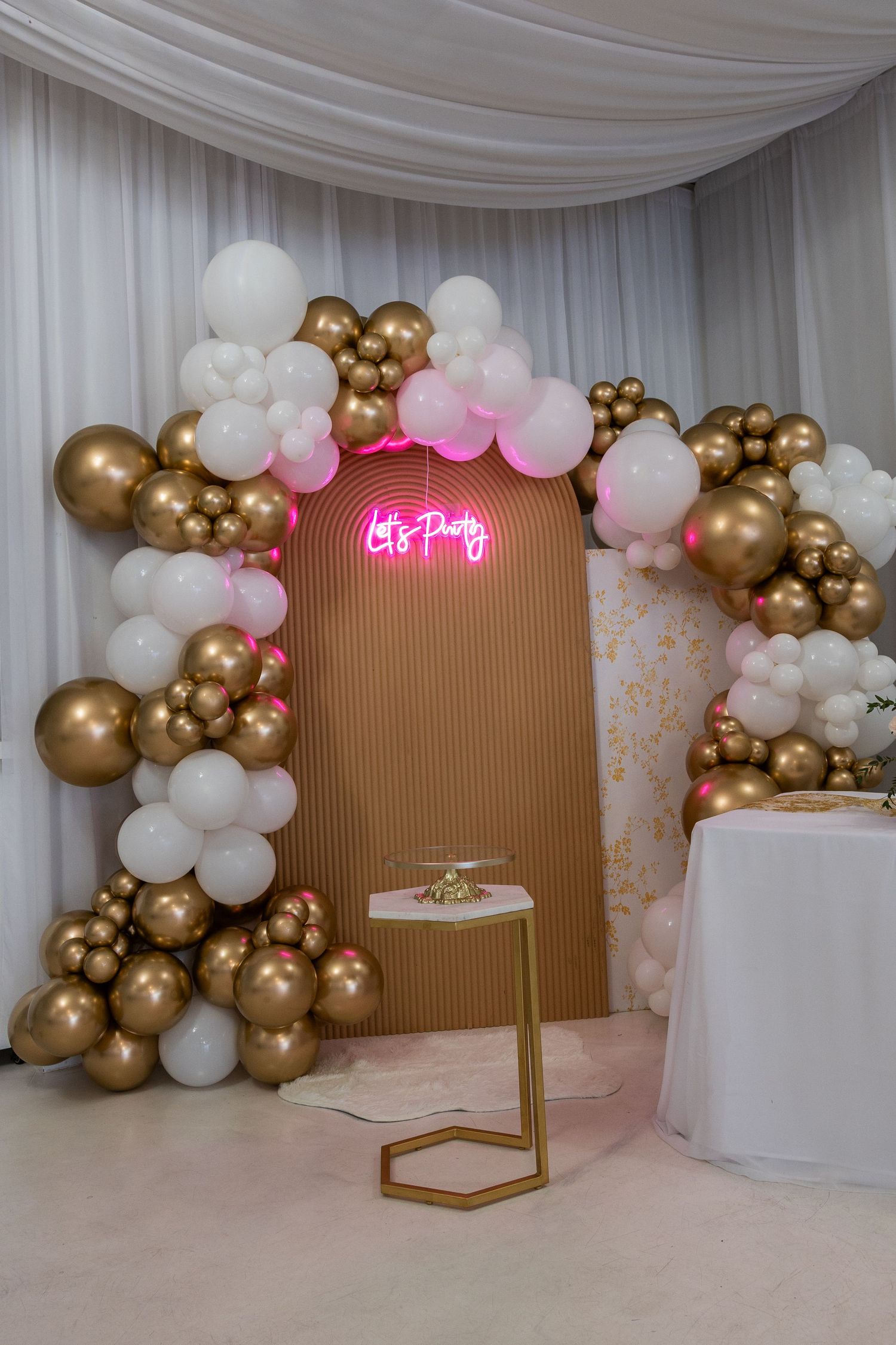 Elegant party backdrop with gold and white balloon garland arch, pink neon "Let's Party" sign, and gold geometric stand.