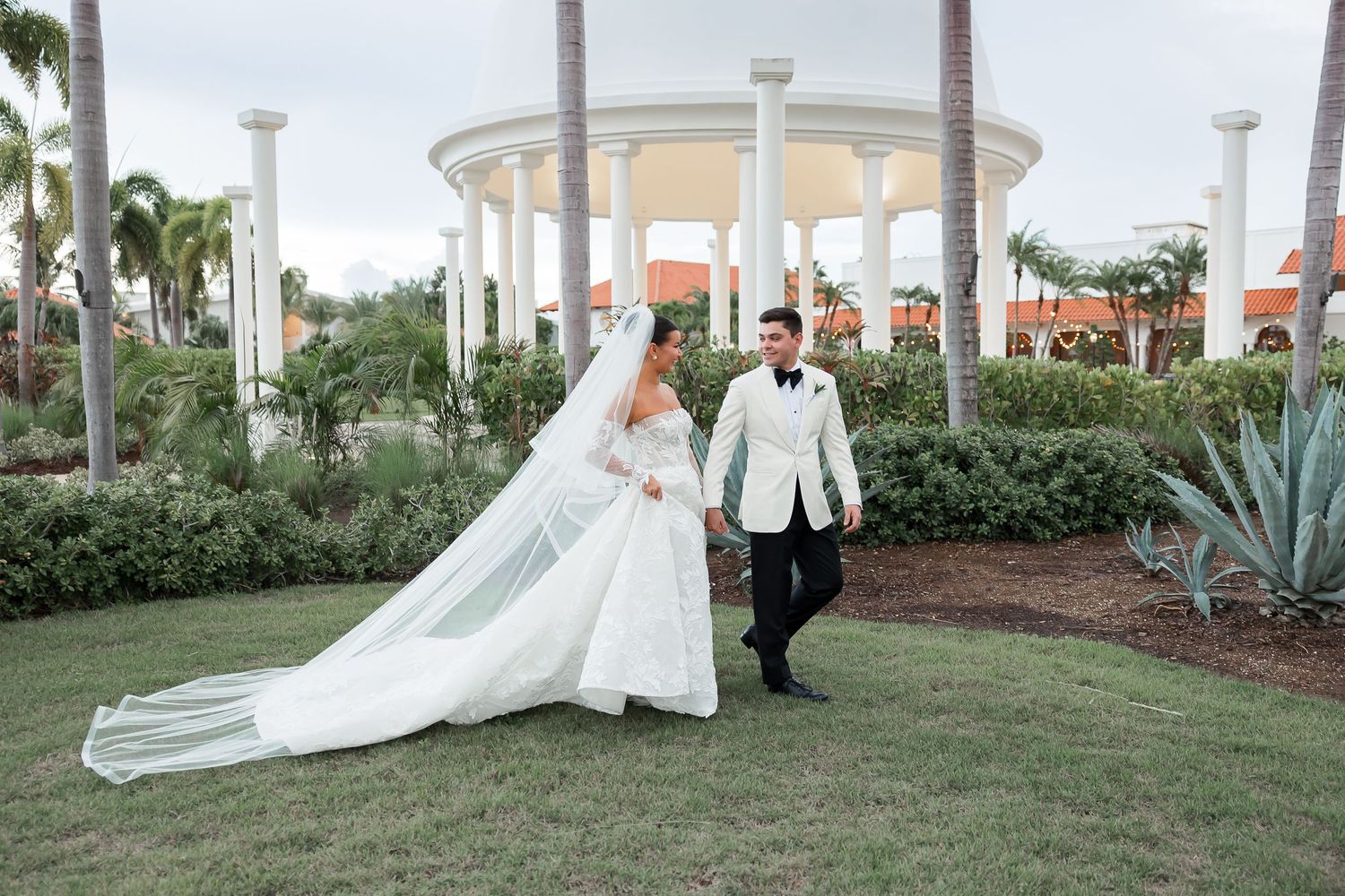 A bride and groom pose for a photo on a grassy lawn with a gazebo in the background.