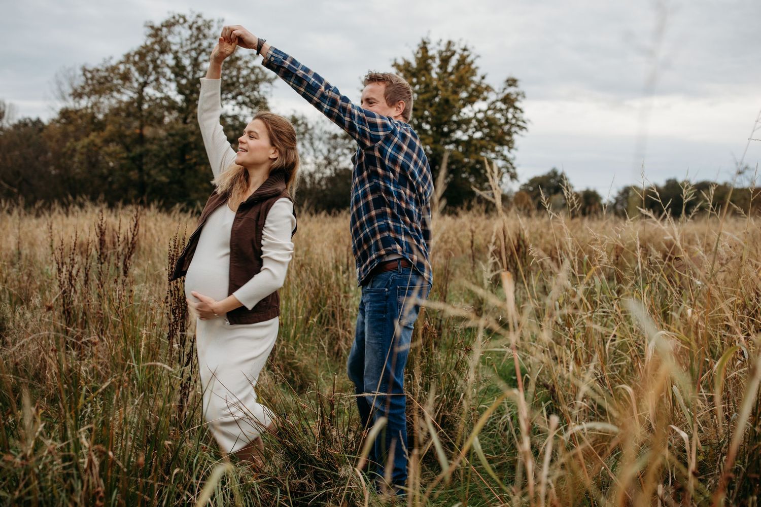 Zwangere vrouw danst met haar man in het veld. Met haar ene hand op haar buk en de andere in die van haar man die haar rond laat draaien.