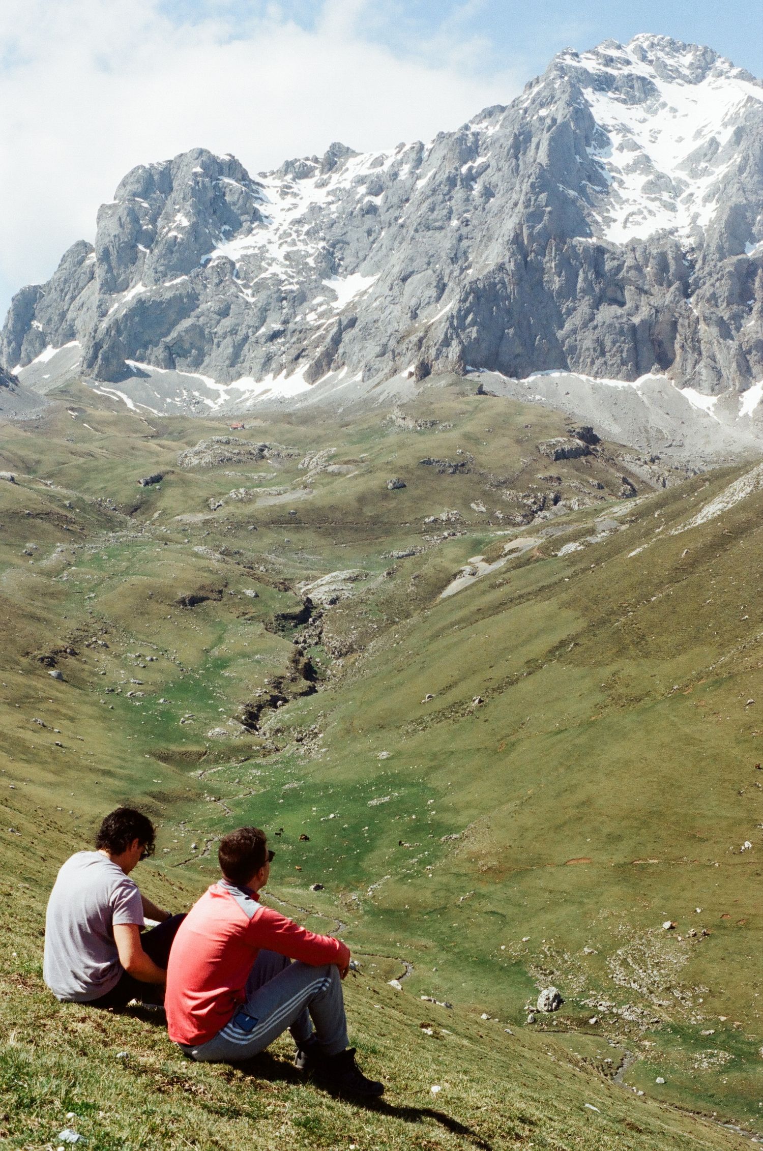 Two people sitting on grassy slope overlooking dramatic mountain landscape.