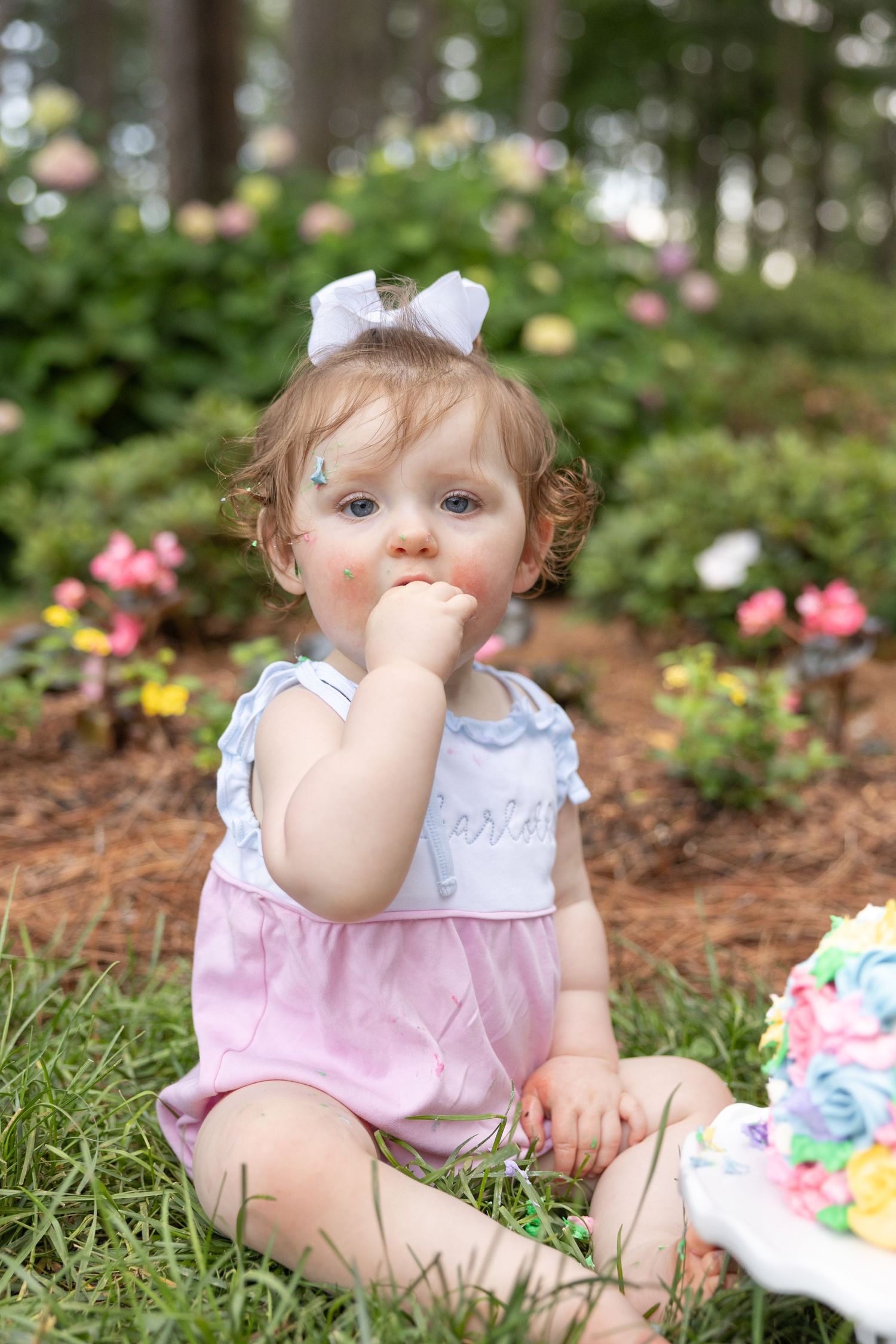 A baby girl with a white bow sits and eats her smash cake at WRAL Azalea Gardens.