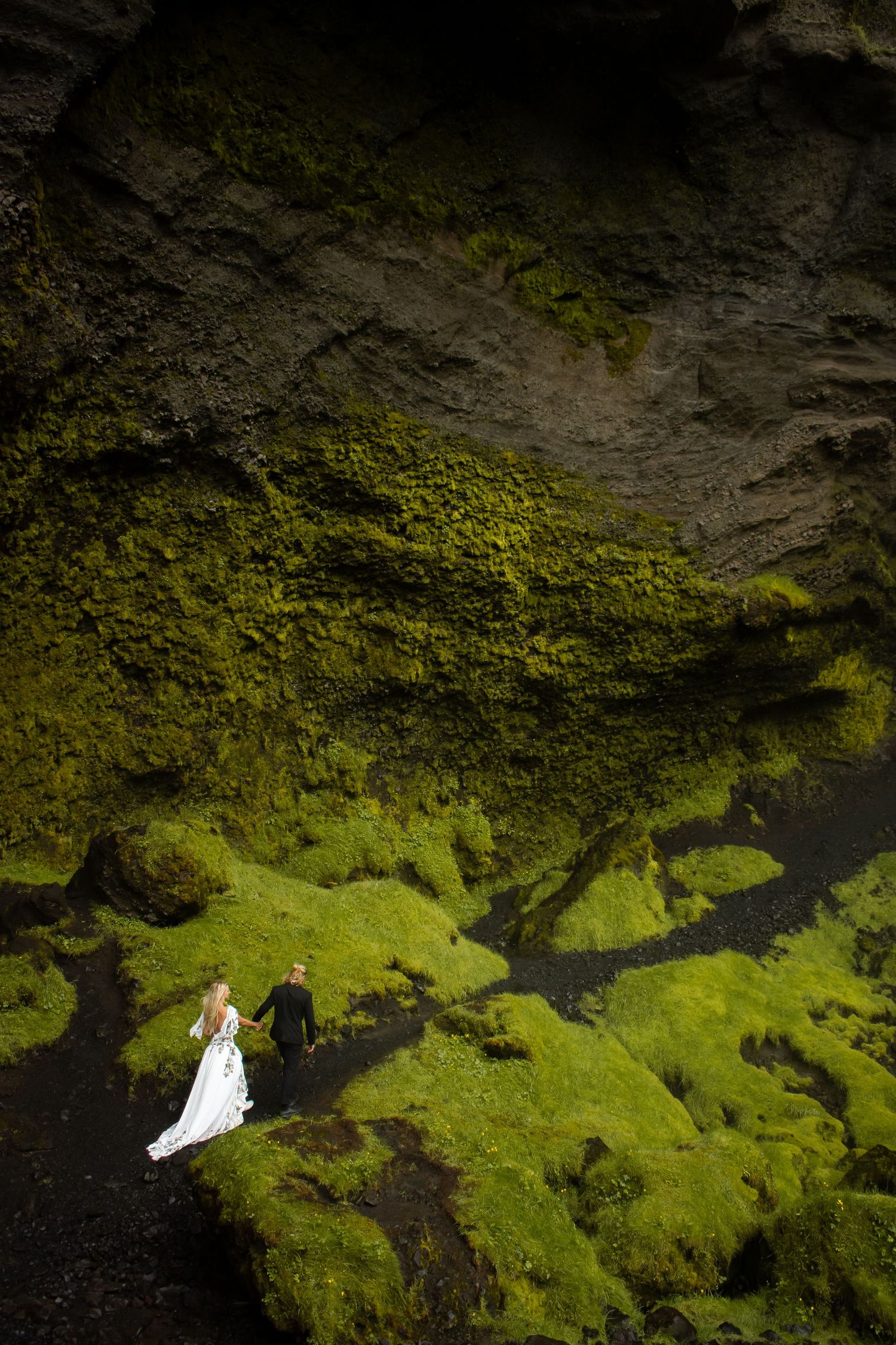 Wedding couple poses on mossy green rocks in dramatic cave setting.
