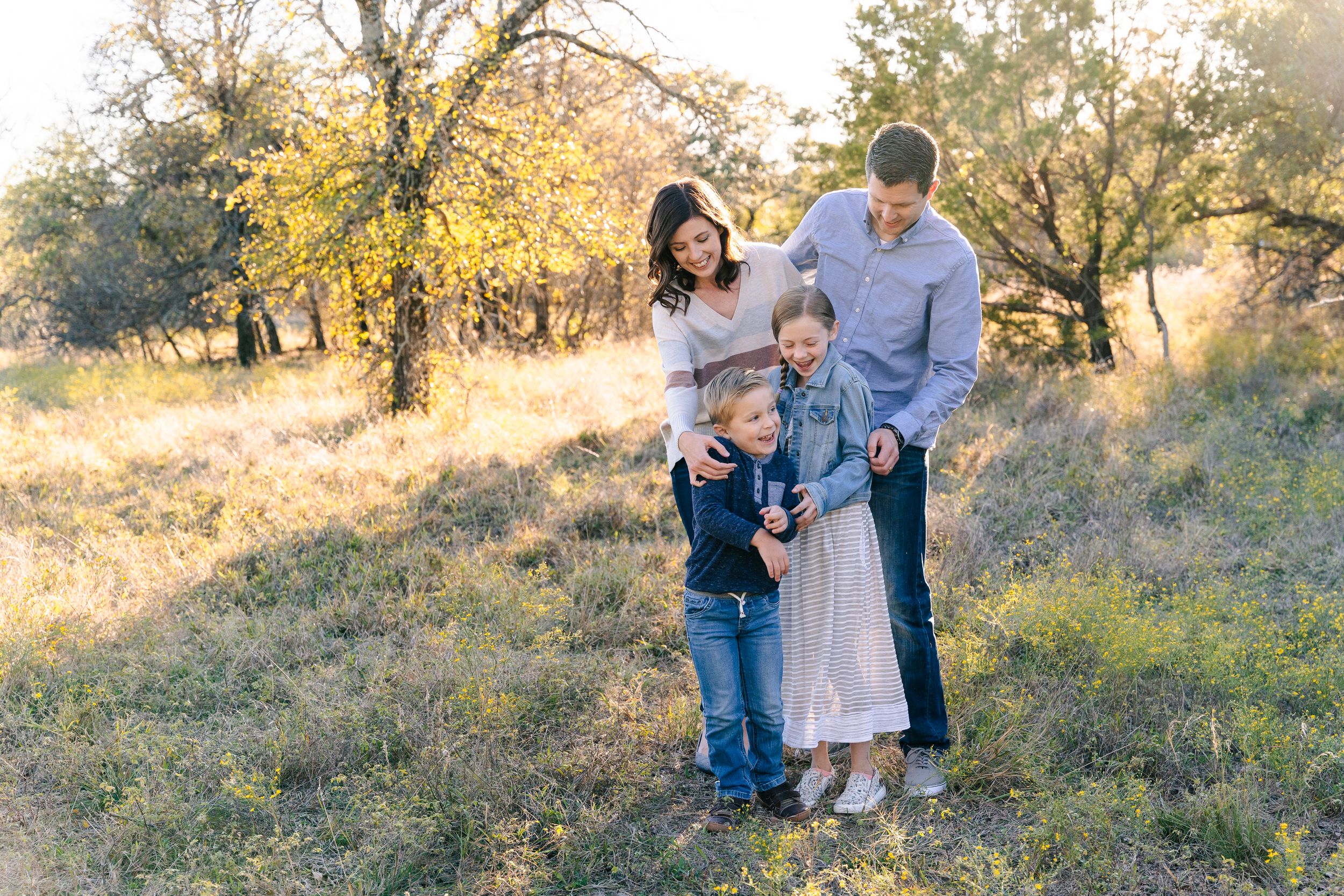 Family Photoshoot in Eagle Mountain Park, TX. Parents tickle their 2 kids in a field with sunlight streaming through the trees behind them.