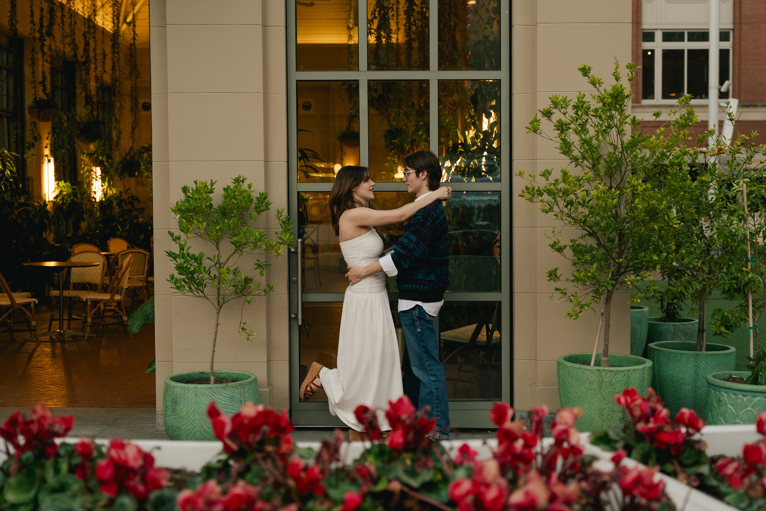 Couples photoshoot in Sundance Square. Girl in a white dress smiles at her fiance as he holds her waist in front of a greenhouse.