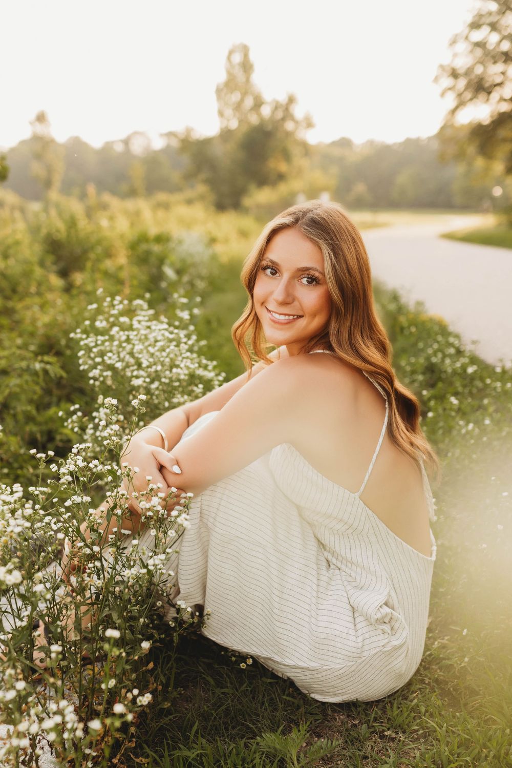 A woman in a white dress sits among blooming wildflowers during golden hour sunset.