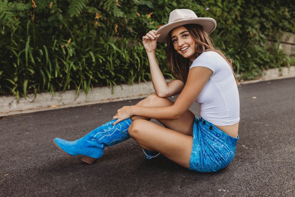 Woman in white top and blue shorts sits casually on asphalt ground while wearing a wide-brimmed hat.