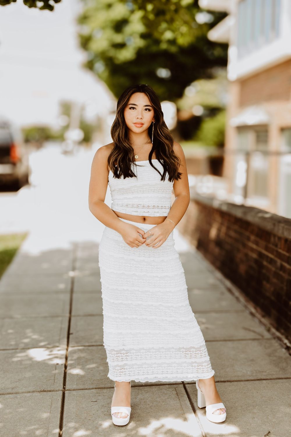 Person in white tiered dress sitting on concrete sidewalk outdoors.