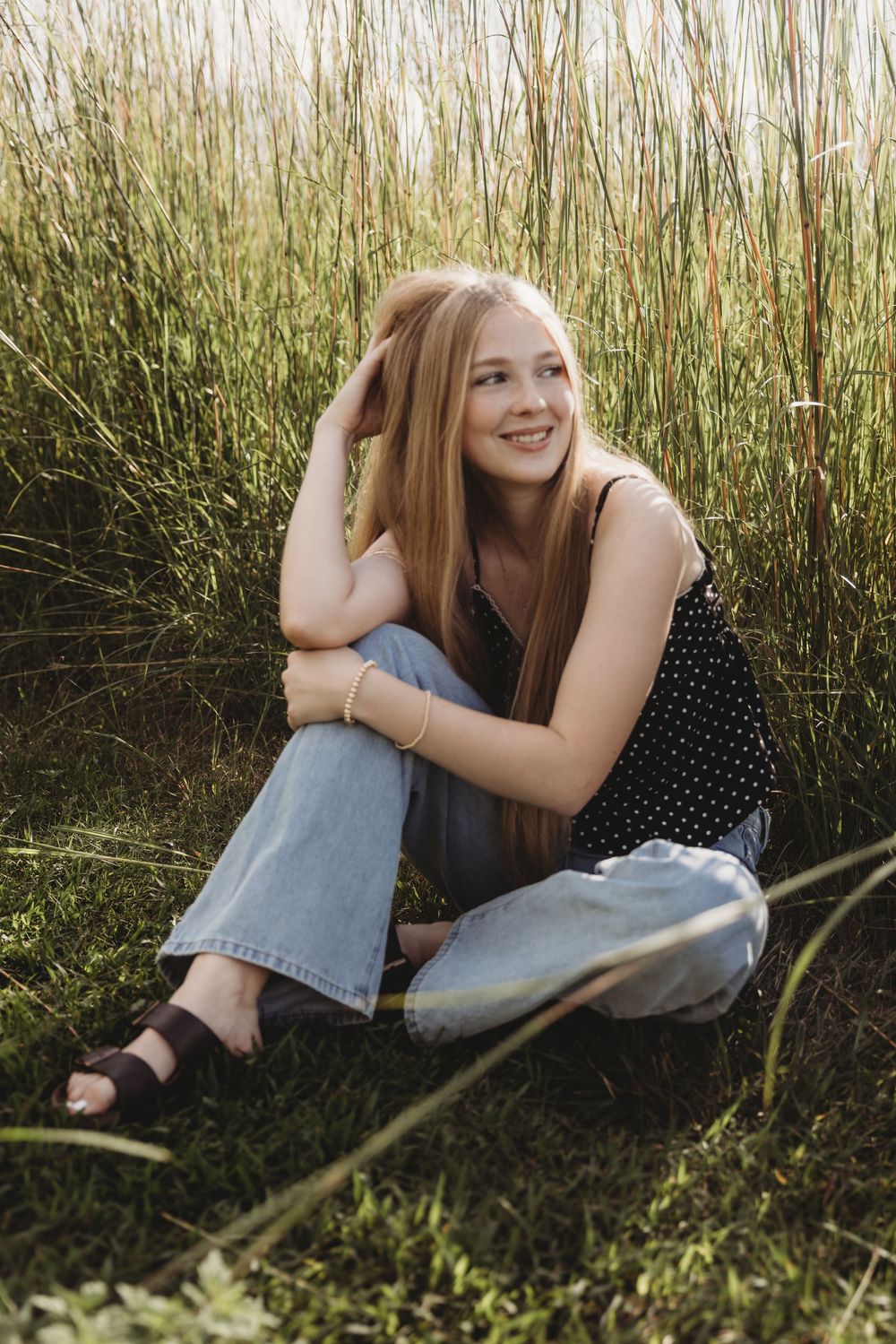 Young woman with long red hair sits smiling in tall green grass, wearing black top, jeans, and sandals.