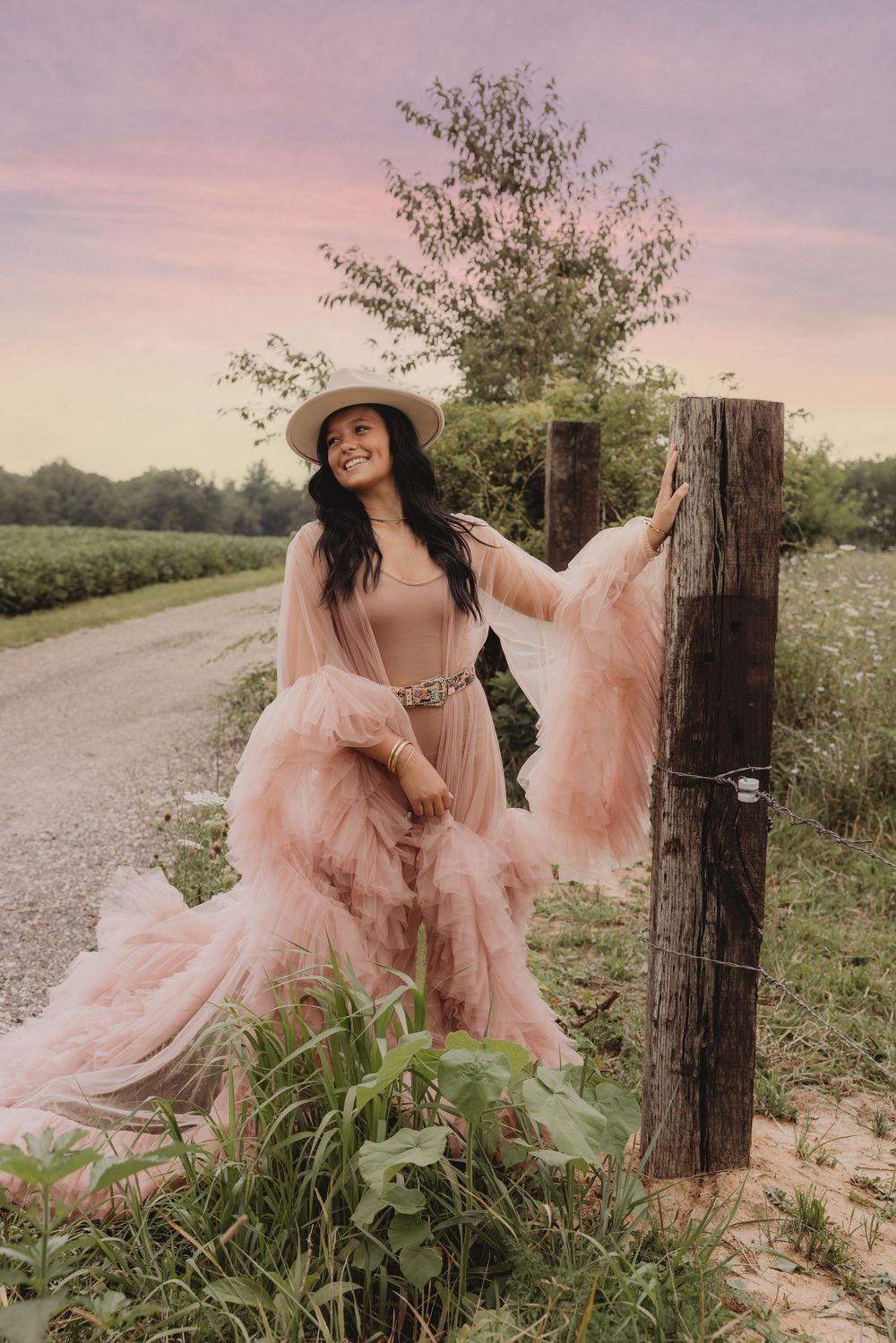 Woman in pink tulle gown and hat poses by rustic fence post at sunset on a country road.