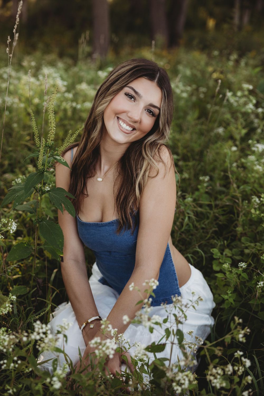Young woman smiling in a wildflower field, wearing a blue top and white skirt, surrounded by lush green plants.