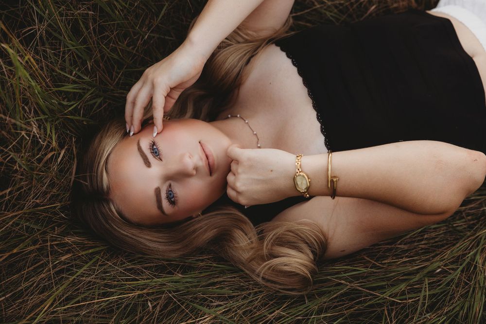 A young blonde woman lying in green grass, wearing a black top and gold bracelet, smiling softly at the camera.