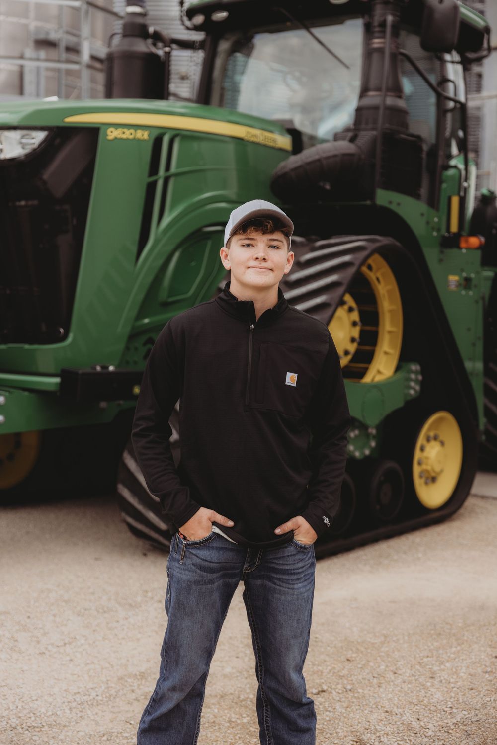 Young man in Carhartt jacket and cap stands confidently before a green John Deere tractor on a farm.