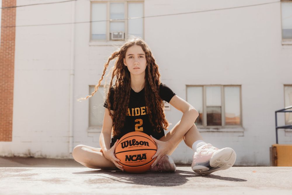 Young female basketball player in Raiders jersey sitting cross-legged on court holding a Wilson NCAA basketball outdoors.