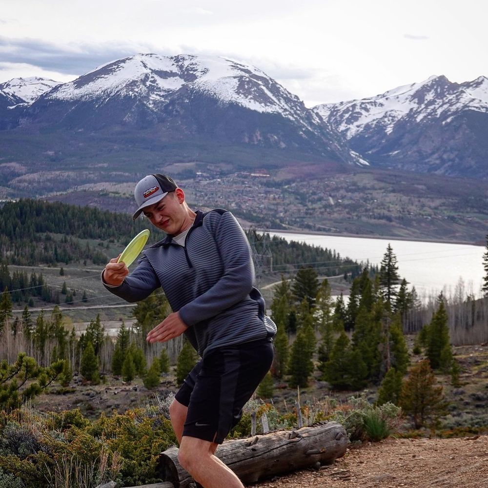 Hiker poses on mountain trail with snow-capped peaks and evergreen forest in scenic mountain landscape backdrop.
