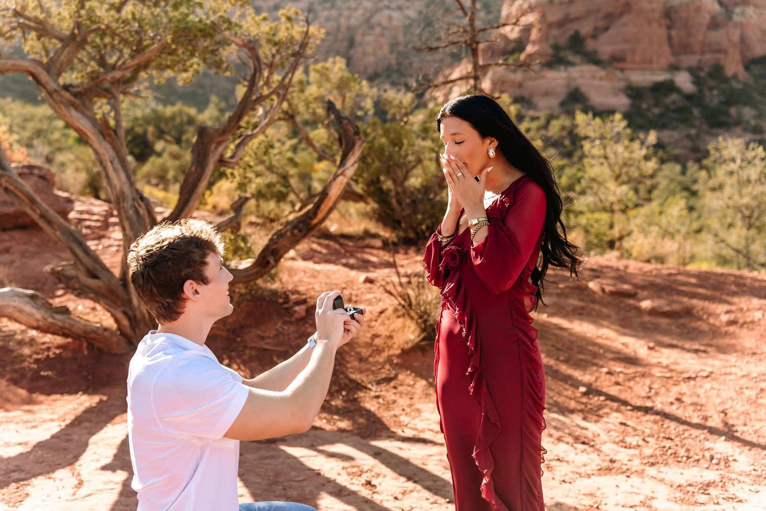 Man proposes to woman on one knee in Sedona Arizona red rock desert landscape, woman covers mouth in surprise.