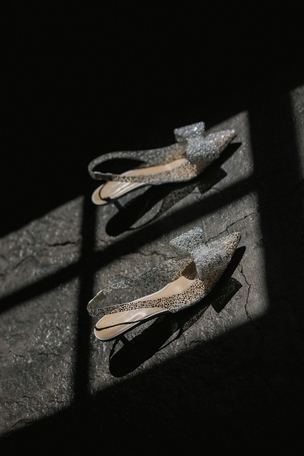 Artistic composition of bridal shoes casting dramatic shadows on dark textured surface.