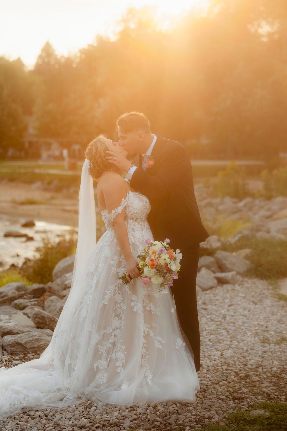 Romantic sunset portrait of couple embracing on rocky beach with bouquet.