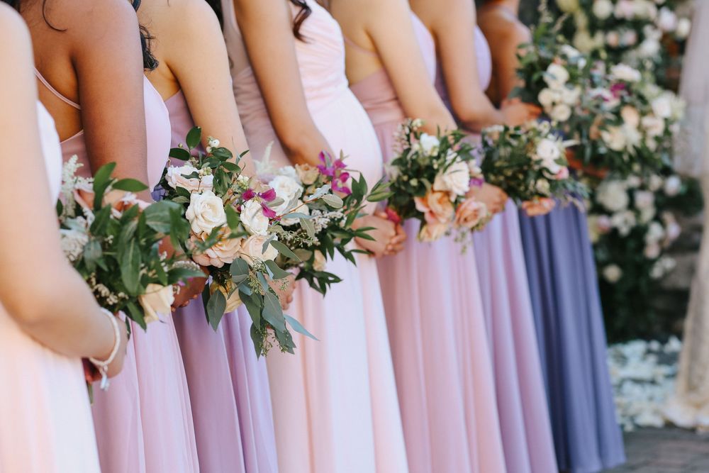 Bridesmaids in pastel pink and purple dresses hold delicate floral bouquets at an elegant wedding ceremony.