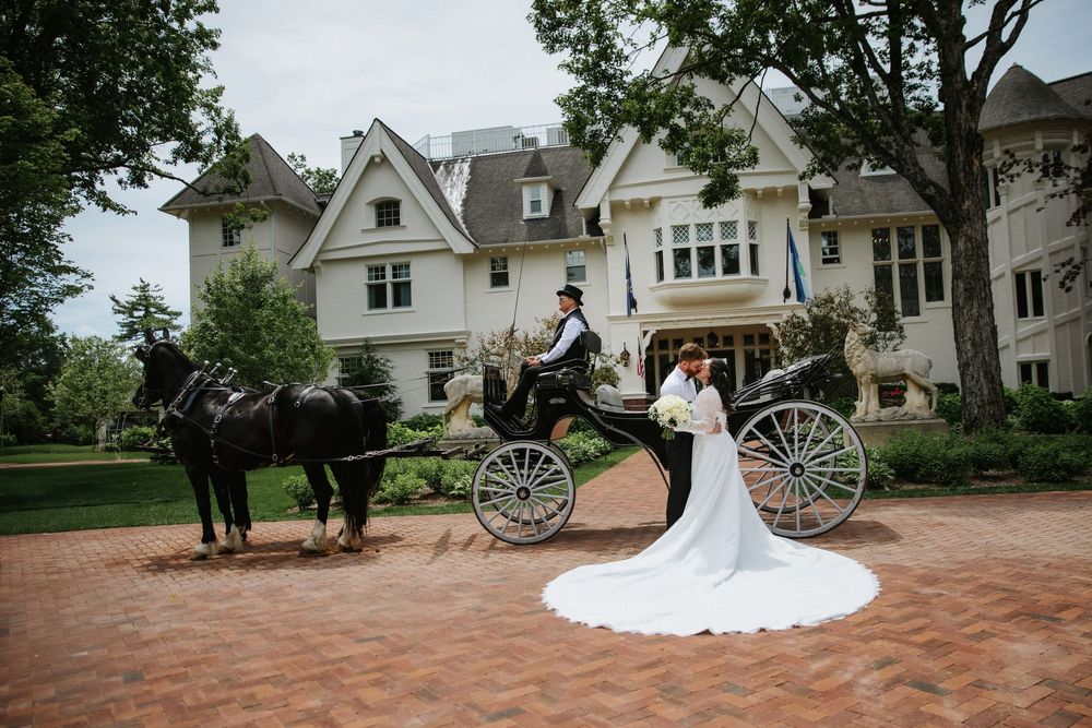 A romantic wedding carriage drawn by horses stands in front of a white mansion on a brick driveway on Mackinac Island Inn at Stonecliffe